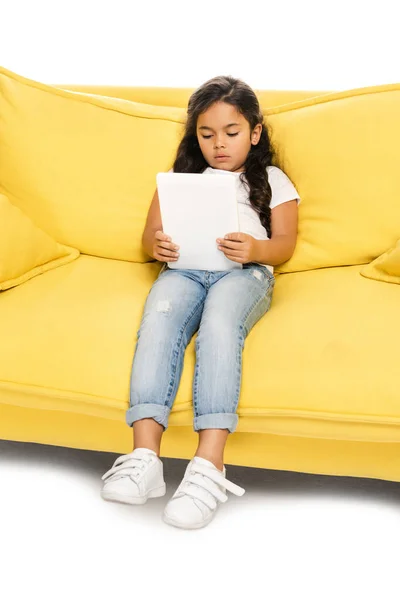 Mignon latin enfant assis sur canapé jaune et en utilisant tablette numérique isolé sur blanc — Photo de stock