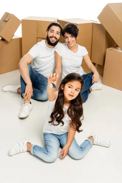 Selective focus of cute latin kid holding keys and sitting near parents and boxes isolated on white — Stock Photo