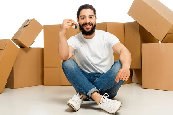 Bearded latin man holding key and sitting with crossed legs near boxes isolated on white — Stock Photo