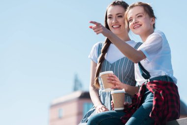 Genç kadın kağıt bardak kahve ile gülümseyen kadın arkadaşına parmak tarafından işaret 