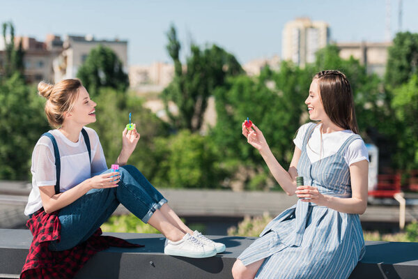 side view of two smiling female friends using bubble blowers at rooftop 
