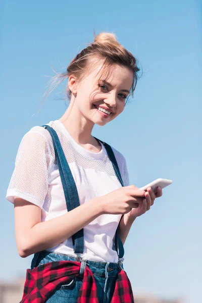 明るい青空に対してスマート フォンを持つ若い女性を笑顔  — 無料ストックフォト
