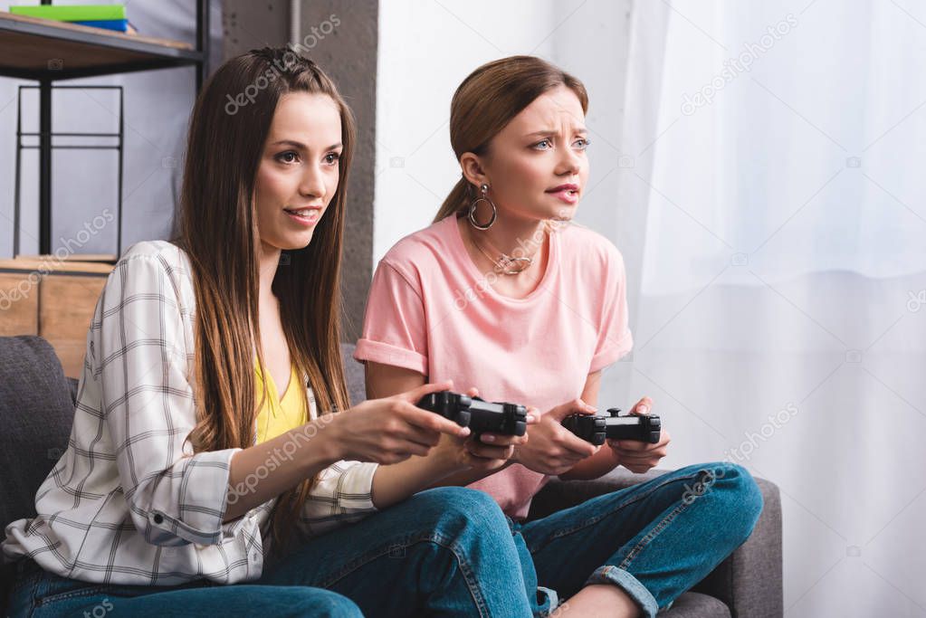 two focused female friends with joysticks in hands playing video game at home