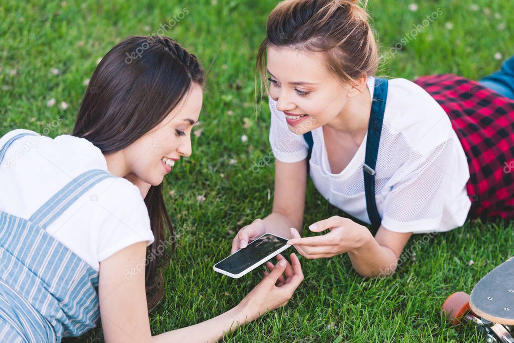 smiling female friends laying on grass with smartphone and skateboard in park 