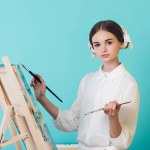Atraente teen menina pintura no cavalete com escova e paleta, isolado no turquesa