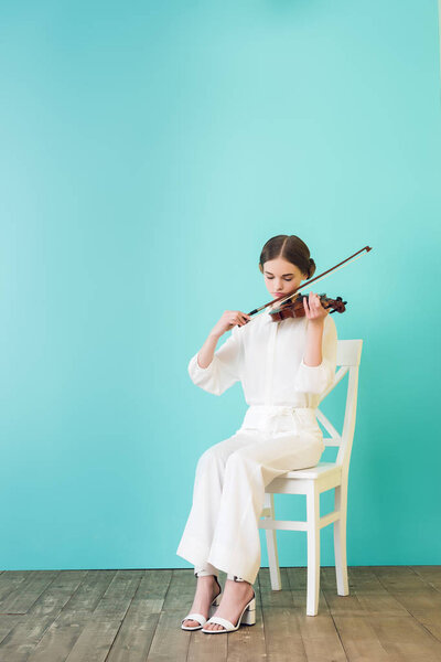 teenager in white outfit playing violin and sitting on chair, on blue