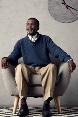 Duvarda büyük saat ile koltukta oturan yakışıklı Afrikalı-Amerikalı adam