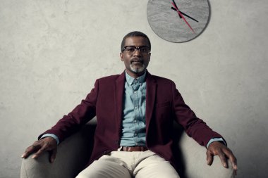 kendine güvenen orta yaşlı Afrikalı-Amerikalı adam duvar saati ile koltukta oturan  