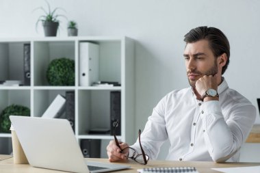 handsome young businessman holding eyeglasses while sitting at workplace and looking at laptop screen clipart