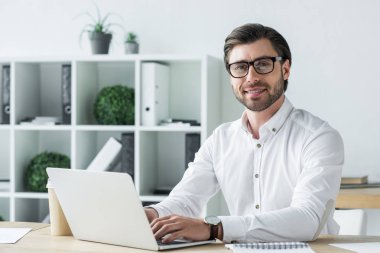 Laptop ile modern ofiste çalışan ve kameraya bakarak mutlu genç işadamı