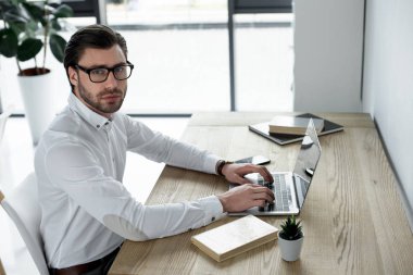 Laptop ile modern ofiste çalışan ve kameraya bakarak kendine güvenen genç işadamı