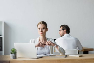 businesswoman sitting at workplace and looking at camera while his colleague working on background clipart