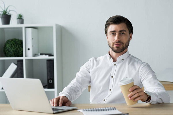 confident young businessman with laptop and coffee to go looking at camera
