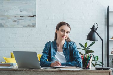 Not alma ve laptop ile evde okurken kameraya gülümseyen genç kız