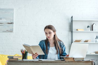 concentrated girl reading book and using laptop while studying at home clipart