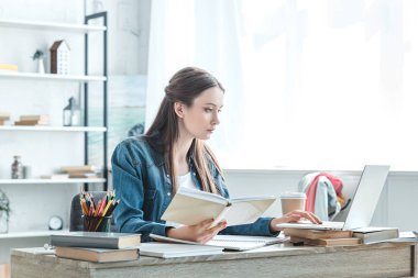 Genç kız holding kitap ve evde eğitim görürken dizüstü kullanarak odaklı