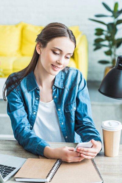 beautiful smiling girl using smartphone while sitting at desk and studying at home