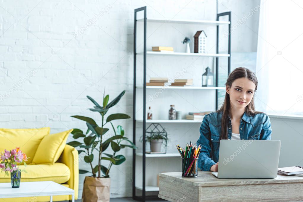 beautiful girl using laptop and smiling at camera while sitting at desk at home