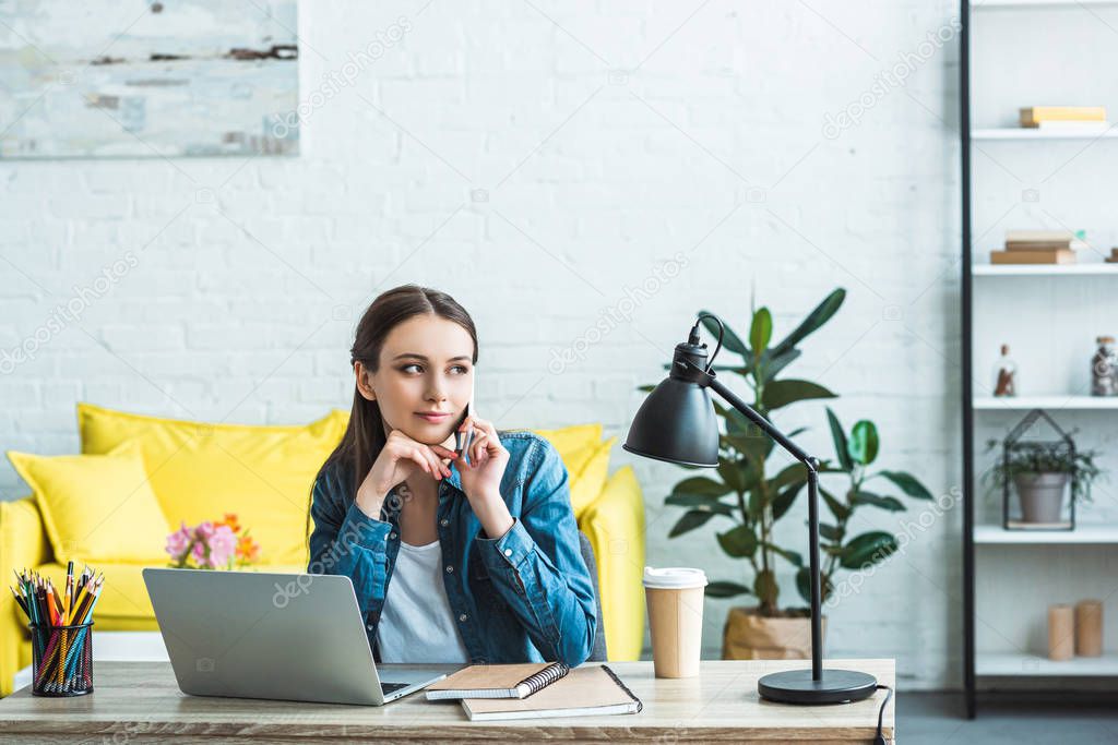 pensive girl talking by smartphone and looking away while studying with laptop and notebooks at home