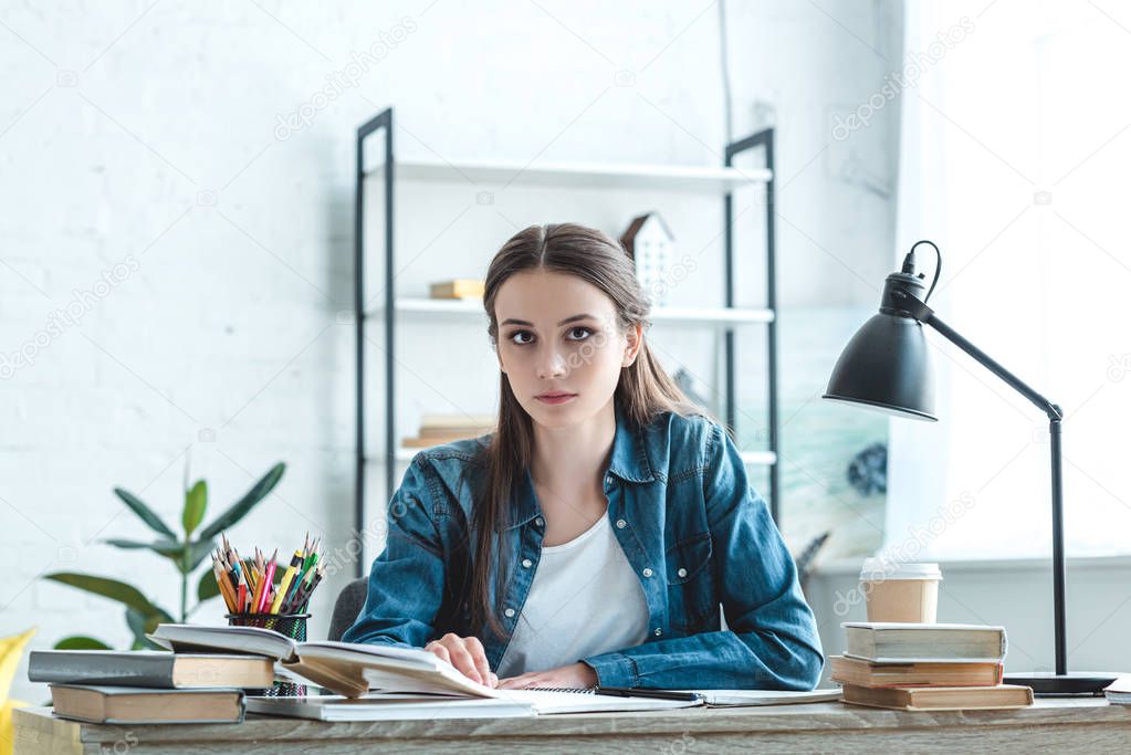 attractive teenage girl sitting at desk and looking at camera while studying at home