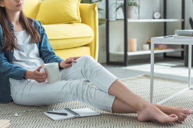 cropped shot of barefoot girl holding cup while sitting on carpet and studying at home clipart