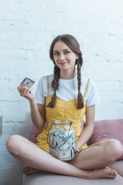 female teenager putting dollar banknote into glass jar for saving while sitting on sofa clipart