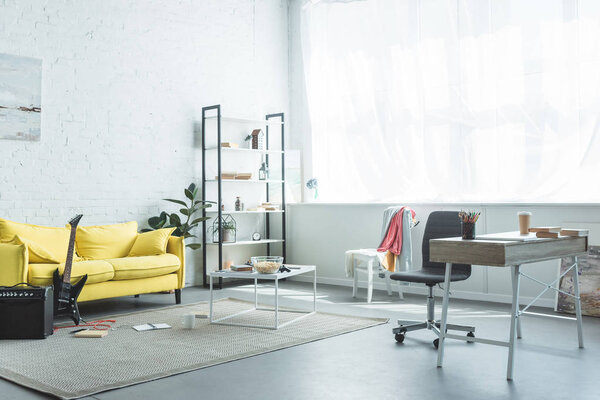 modern room interior with coffee to go and books on wooden desk and electric guitar near yellow couch 