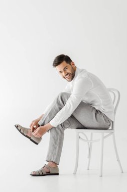 smiling man in linen clothes sitting on chair and putting on sandals isolated on grey background 