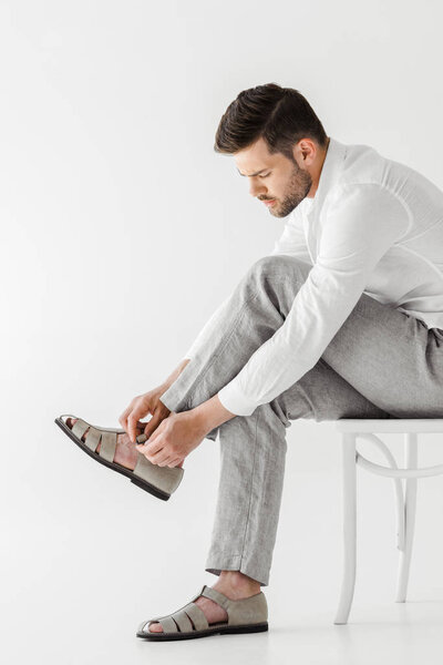 side view of man in linen clothes sitting on chair and putting on sandals isolated on grey background 