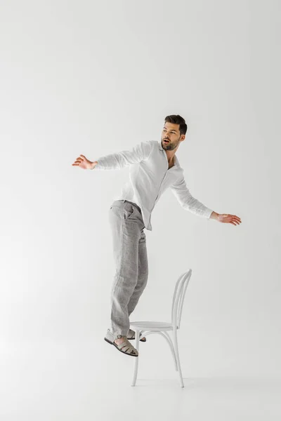 灰色の背景に分離された椅子の上に立って広い腕とリネンの服の男 — ストック写真