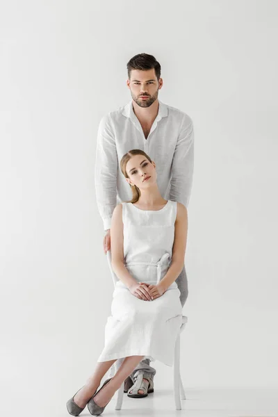 魅力的な女性は彼氏に孤立した灰色の背景の後ろに立っている間椅子に座ってリネンの白いドレスで — ストック写真