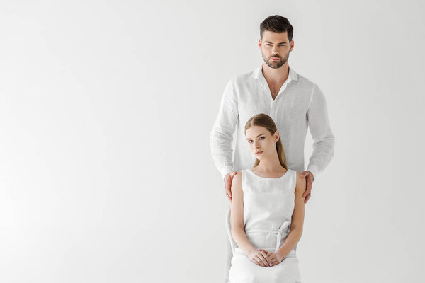 beautiful woman in linen white dress sitting on chair while her boyfriend standing behind isolated on grey background 
