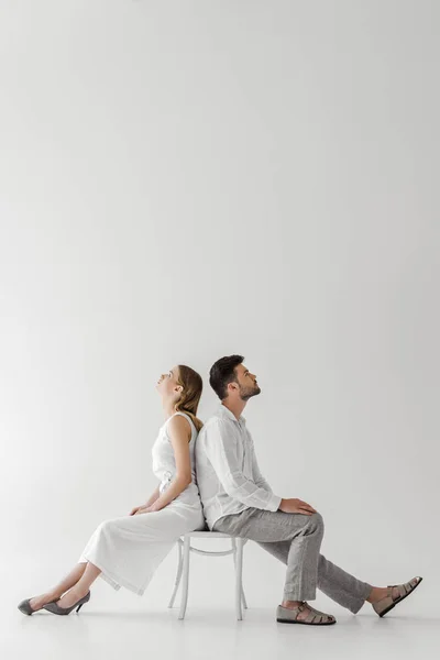 灰色の背景に分離されて椅子に背中合わせに座っているリネンの服のモデルの若いカップルの側面図 — ストック写真