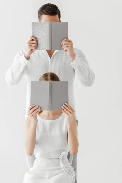 couple in linen clothes covering faces by books isolated on grey background