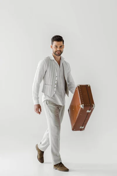 灰色の背景に分離されたヴィンテージのスーツケースを運ぶリネンの服の流行の男性旅行者の笑顔 — ストック写真