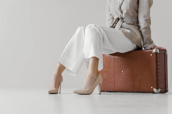 cropped image of stylish woman in linen jacket sitting on vintage suitcase isolated on grey background 