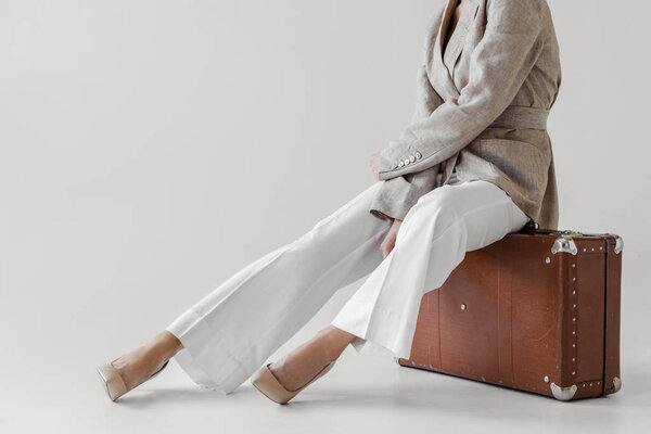 cropped image of stylish woman in linen jacket sitting on vintage suitcase isolated on grey background 