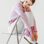 Stylische junge Frau in rosa Kapuzenpulli sitzt auf Stuhl auf weiß Stylische junge Frau in rosa Kapuzenpulli sitzt auf Stuhl auf weiß