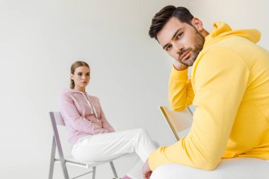 stylish young male and female models in pink and yellow hoodies sitting on chairs isolated on white clipart