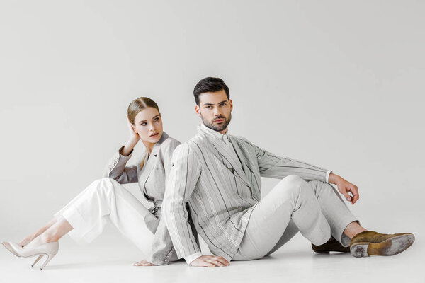 пара привлекательных моделей в винтажной одежде, сидящих на полу и склоняющихся спиной к спине на белом
