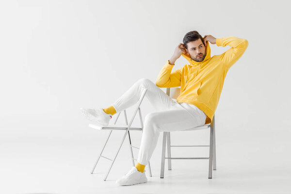 handsome young man in yellow hoodie putting on hood while sitting on chairs on white