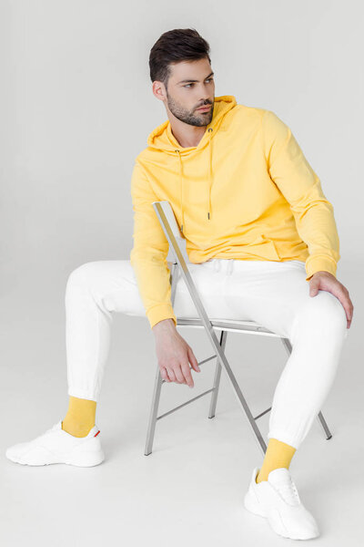 stylish young man in yellow hoodie and white pants sitting on chair and looking away on white