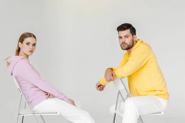 side view of young male and female models in pink and yellow hoodies sitting on chairs and looking at camera on white