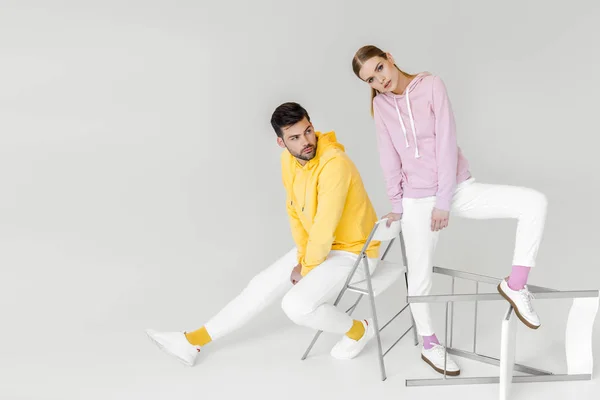 stylish young male and female models in colorful hoodies sitting on chairs on white