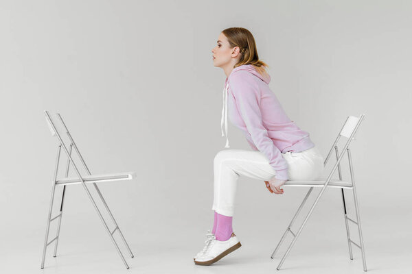 side view of stylish young woman sitting on chair in front of another empty chair on white