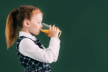 side view of adorable little schoolgirl drinking orange juice while standing near chalkboard clipart