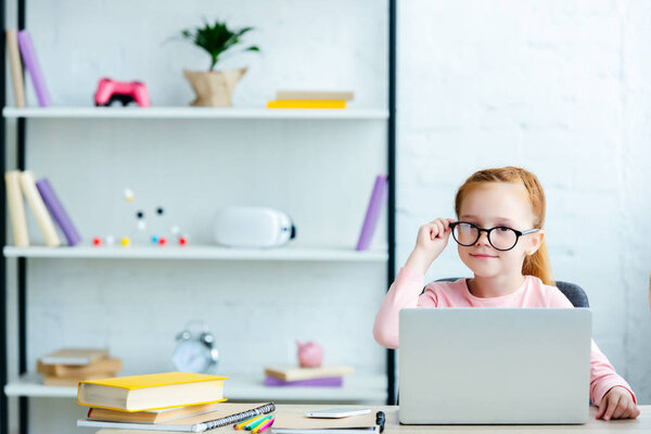 adorable red haired schoolgirl in eyeglasses using laptop and smiling at camera while studying at home