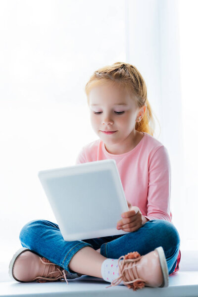 beautiful smiling child using digital tablet while sitting on windowsill  