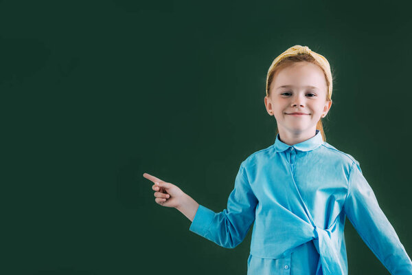 beautiful red haired schoolgirl pointing at blank chalkboard and smiling at camera