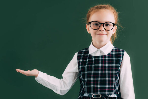 adorable little schoolgirl in eyeglasses showing blank chalkboard and smiling at camera 